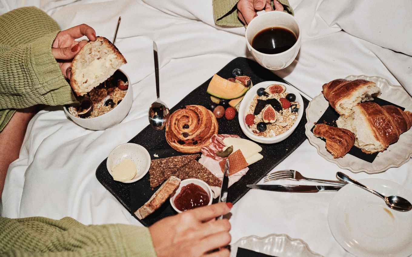 A breakfast spread on a bed, including fresh fruit, pastries, granola, cheese, bread, and coffee, with hands reaching in.